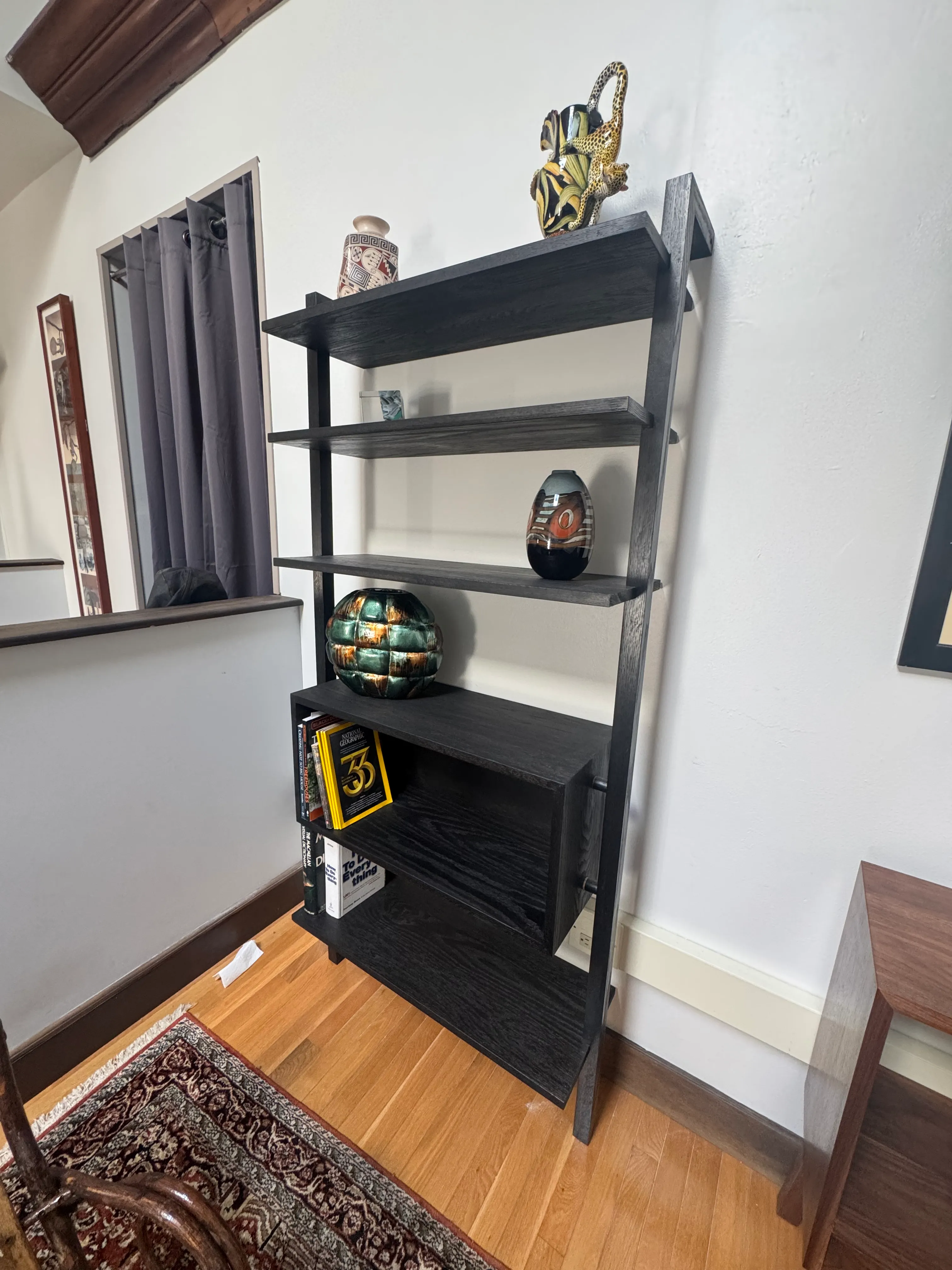 A blackened red oak leaning bookshelf holding books and pottery against a white wall.