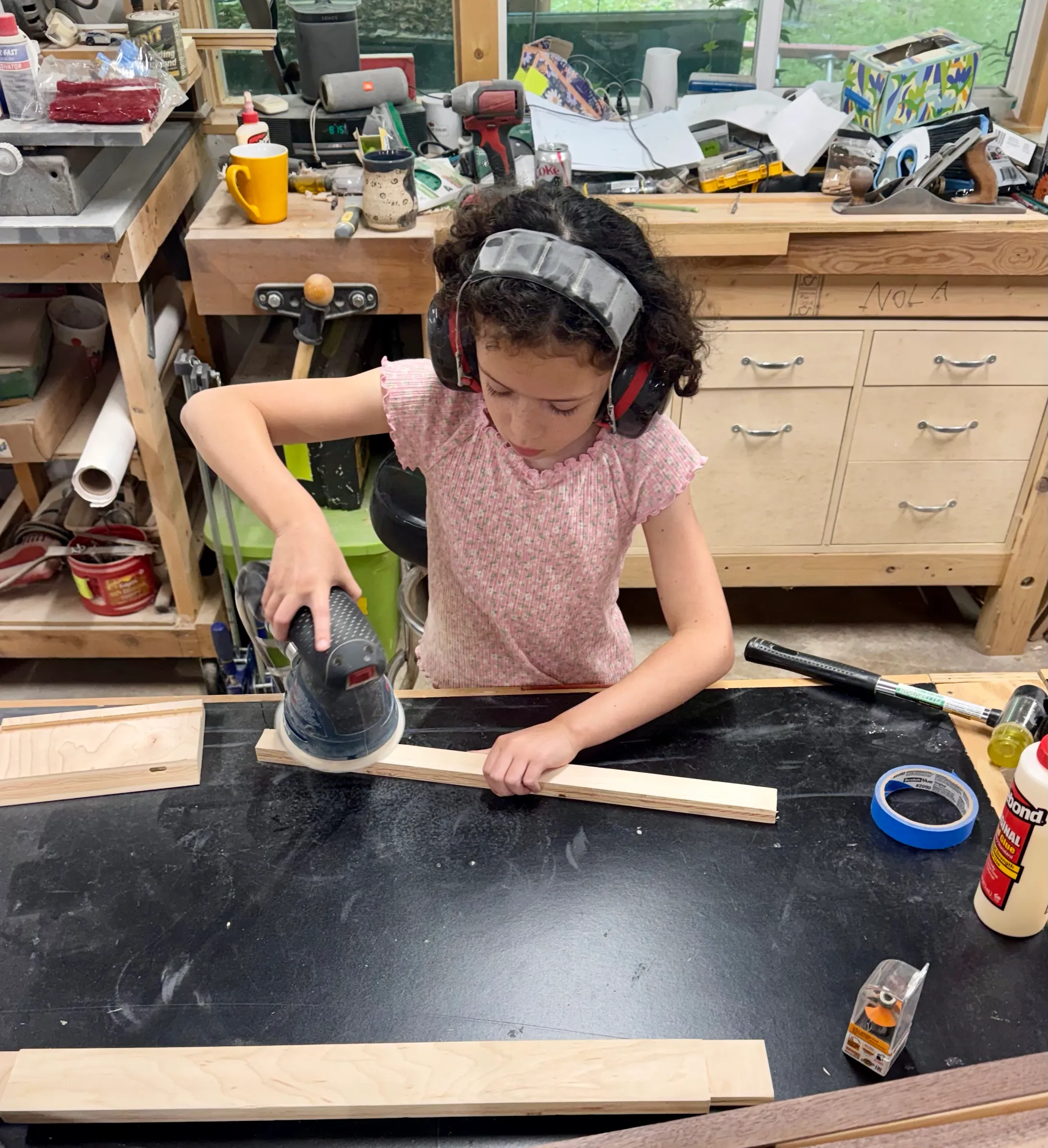 Nola, a young child in a pink shirt and ear protection, running a random-orbit sander along a board on the workbench.