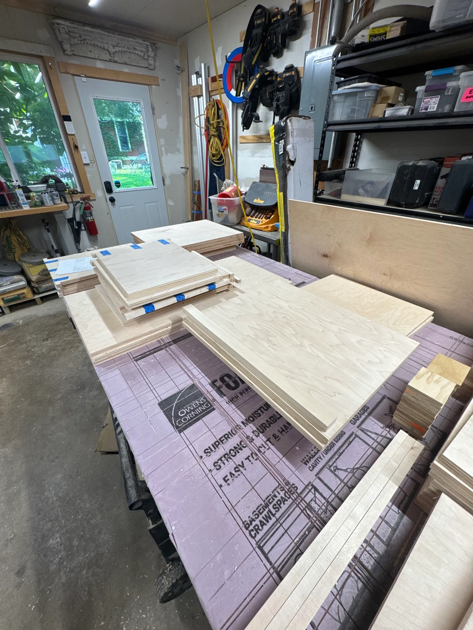 Maple plywood cut to size for the cubby boxes, stacked and labeled with blue tape on the shop assembly table.