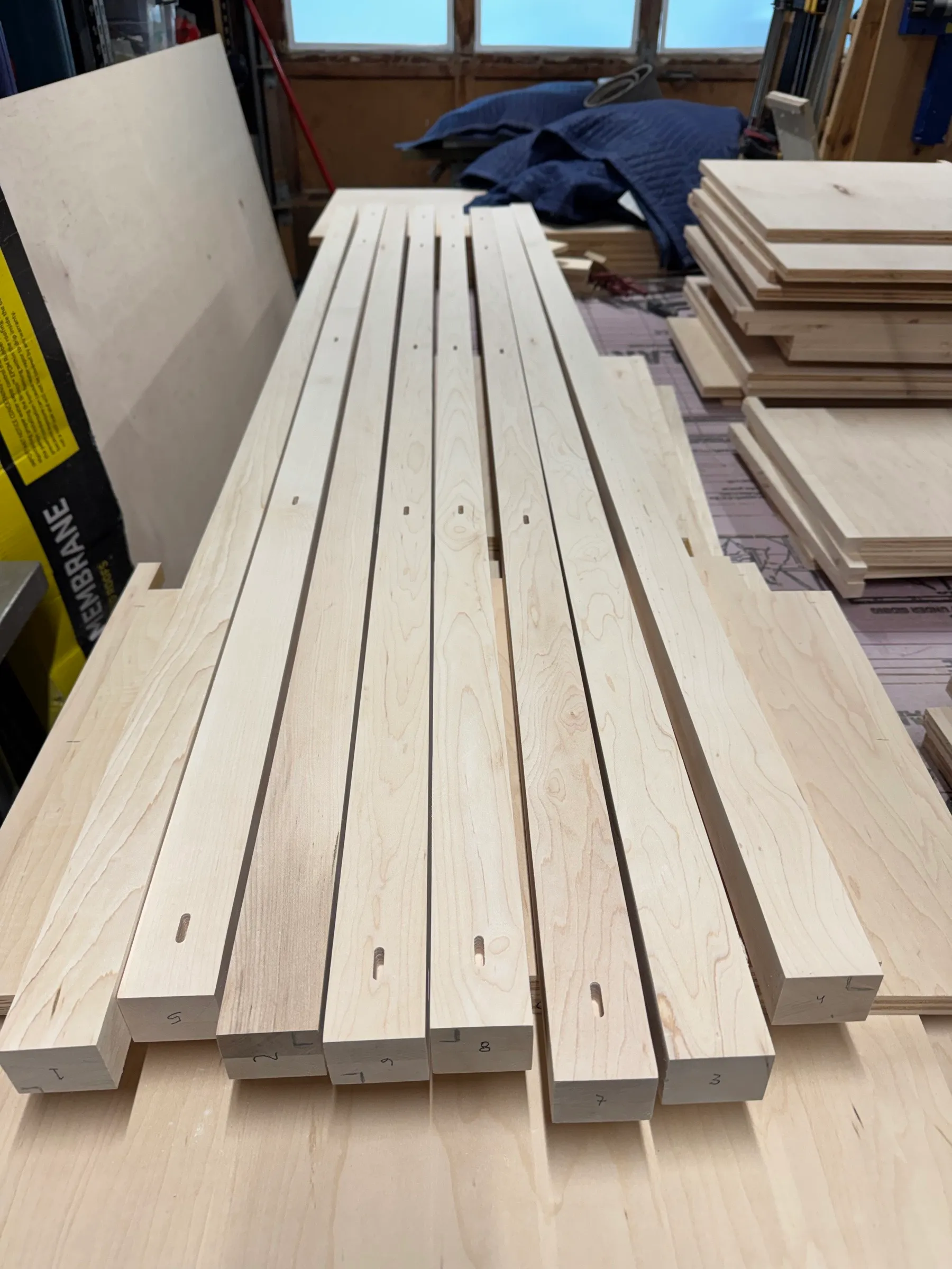 Numbered maple frame members laid side by side, each showing a routed mortise, with plywood box parts stacked behind.