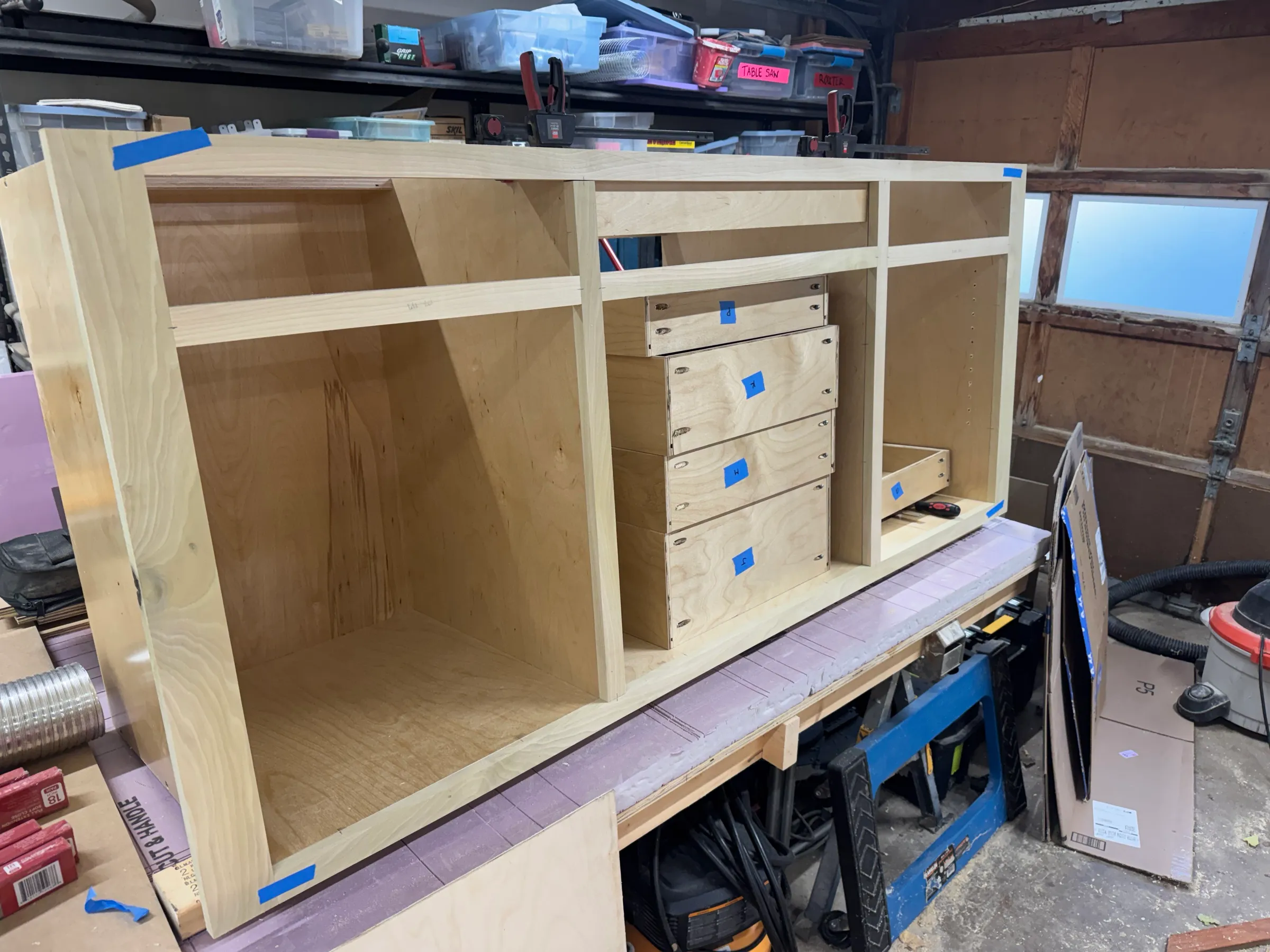 Plywood vanity carcass on the bench with a stack of labeled drawer boxes waiting in the center bay.