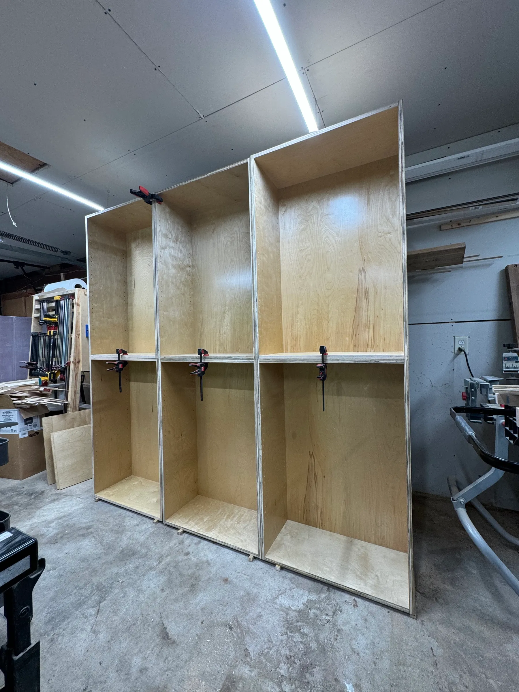 Two plywood shelf carcasses clamped together on the shop floor, empty adjustable shelves inside.