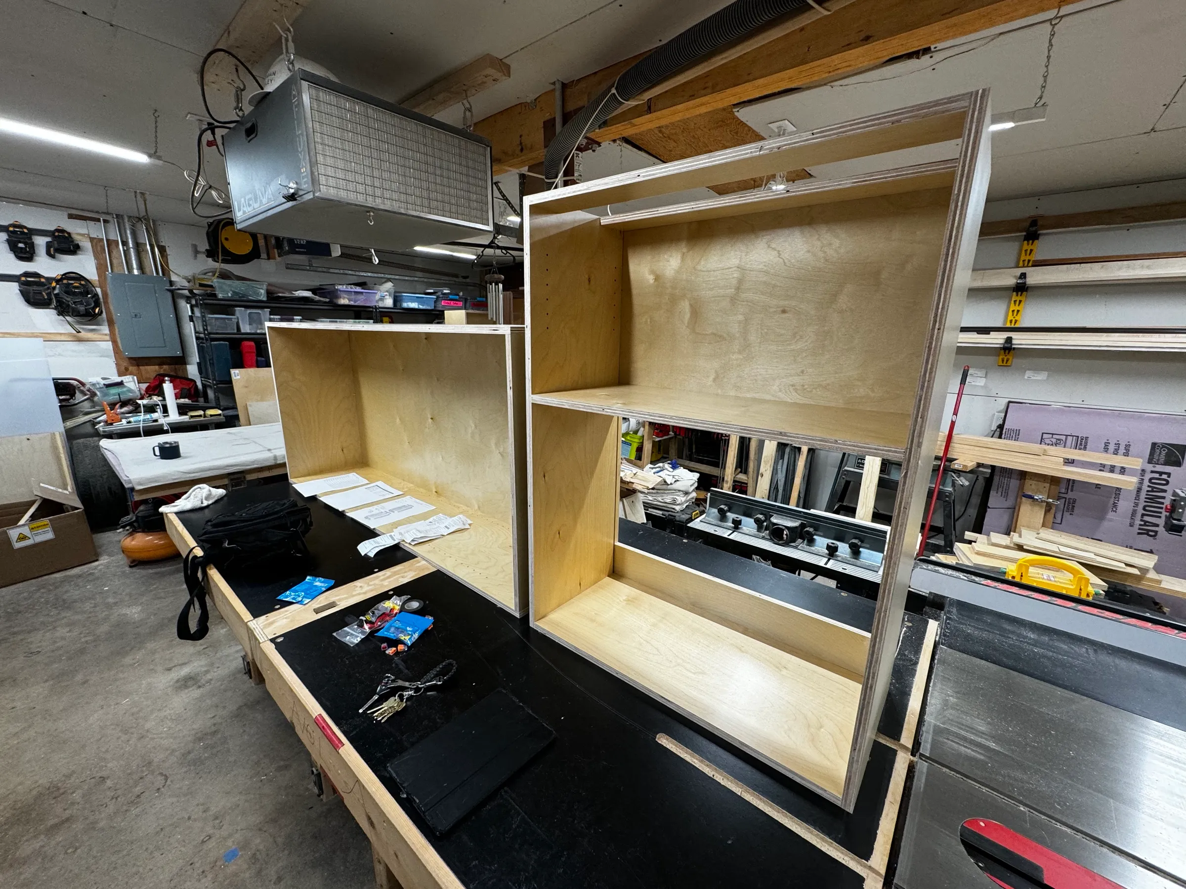 Two plywood carcasses on the shop bench, one shelf still loose, with the dust collector running overhead.