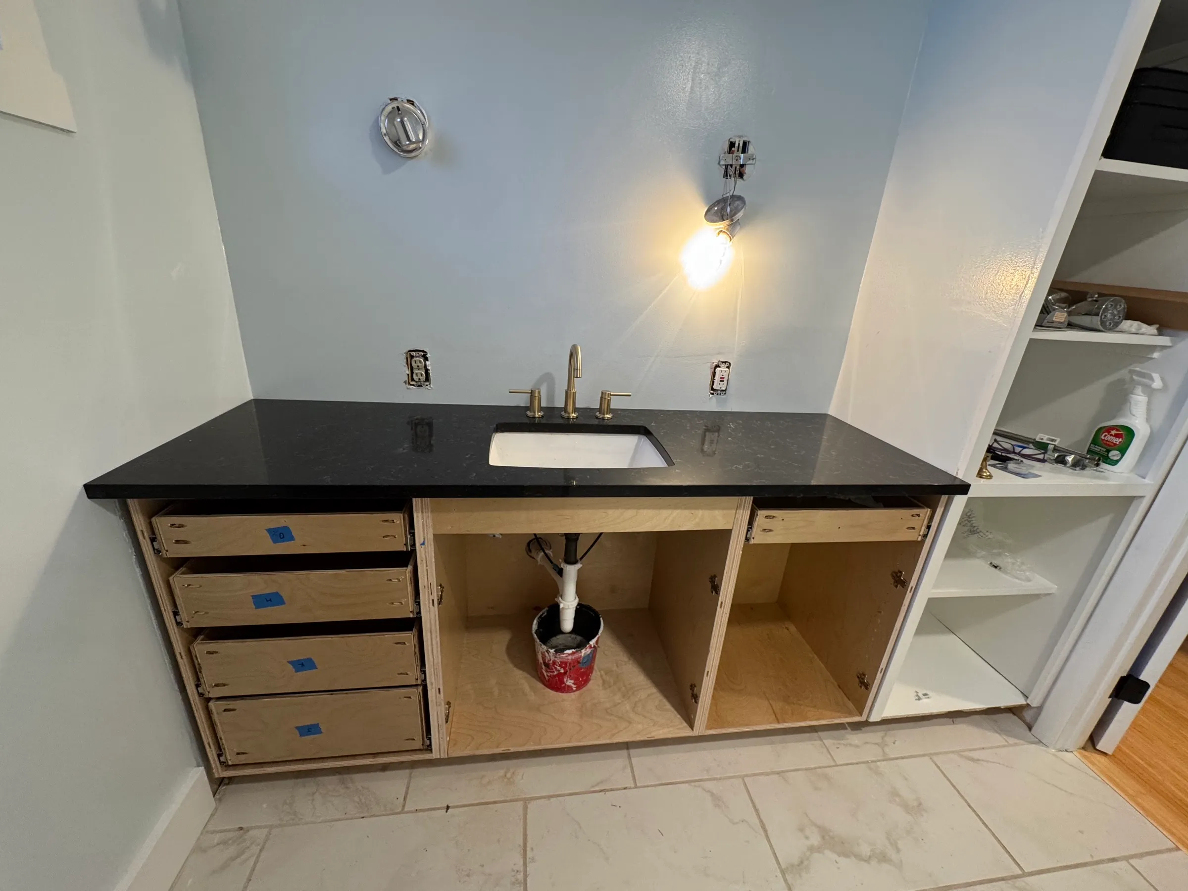 The vanity part-installed in the bathroom, honed black stone top and brushed brass faucet set, drawer boxes open below.