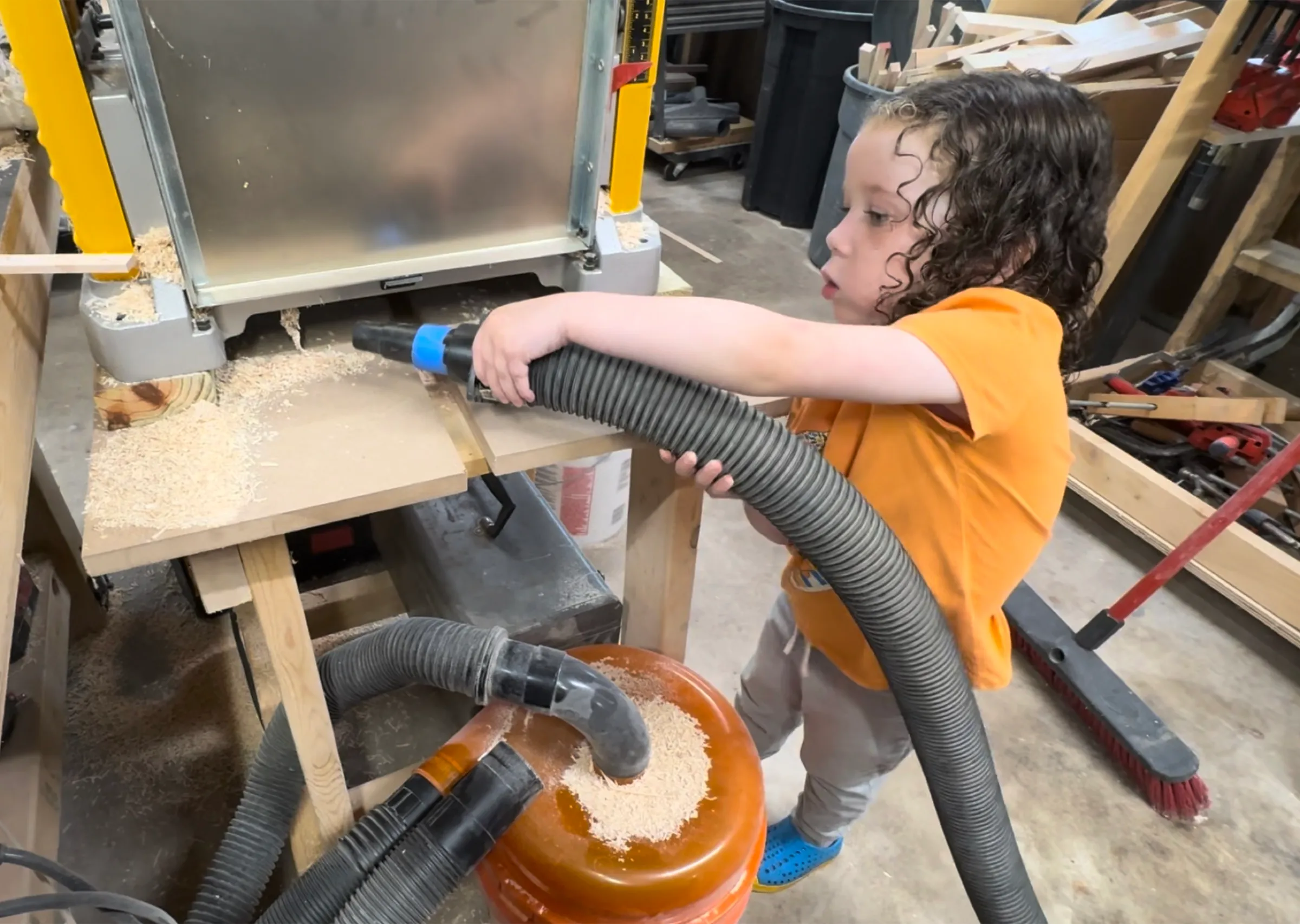 Sebelle, a young child in an orange shirt, holding a shop-vac hose to vacuum sawdust off a workbench in the shop.